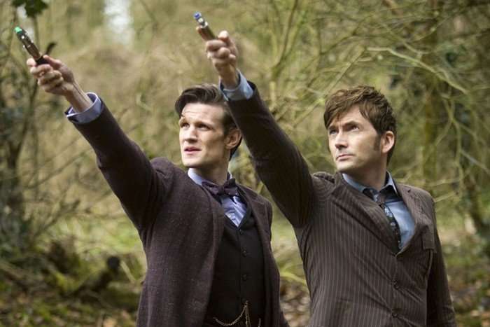 Matt Smith and David Tennant as The Doctor point sonic screwdrivers in the air as they look above at something.