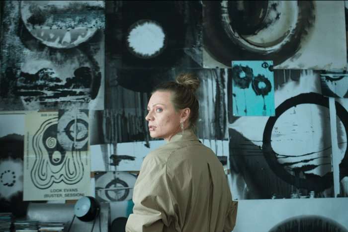 netflix harlan A woman with her hair tied in a loose bun stands in front of a wall covered in abstract black-and-white artwork and posters. Wearing a beige jacket, she glances back over her shoulder with a wary, unsettled expression. The stark monochrome designs surrounding her create an artistic yet ominous atmosphere, hinting at mystery and intrigue.
