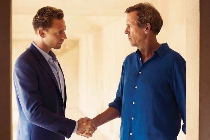 night-manager Tom Hiddleston, dressed in a navy suit, and Hugh Laurie, dressed in a blue shirt, shake hands in a warmly lit corridor in a scene from The Night Manager