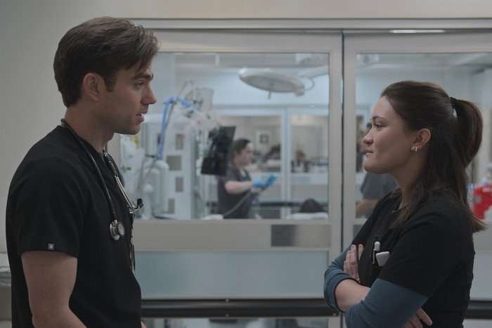 Patrick Ball and Isa Briones in The Pitt, wearing blue scrubs, stood in front of a window into a hospital room and facing each other.