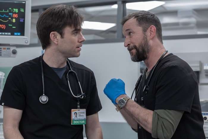 Noah Wyle and Patrick Ball in The Pitt, both wearing blue scrubs and talking with one another intensely.
