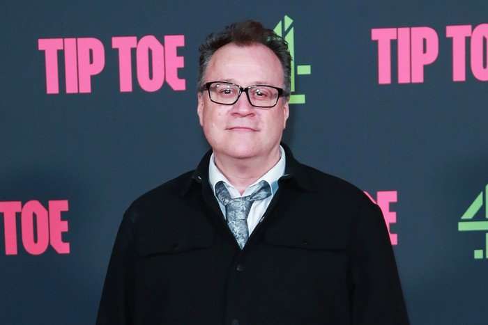 Russell T Davies photographed wearing a white shirt, blue tie and black jacket at the premiere of Channel 4's Tip Toe, standing in front of a board with the show's logo on it