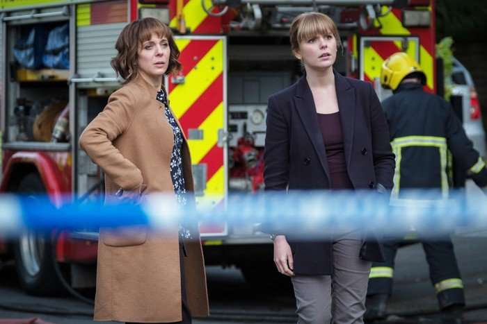 Safe Two women stand behind a police cordon, looking concerned as they watch an incident unfold. Behind them, a fire engine and a firefighter in protective gear are visible, suggesting an ongoing emergency response.
