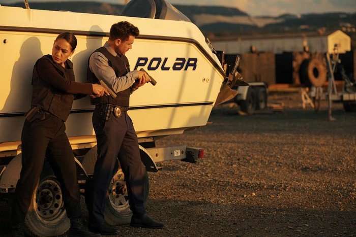 Jos&eacute;phine Jobert and Allan Hawco in Saint-Pierre hiding behind a boat, with their guns ready.