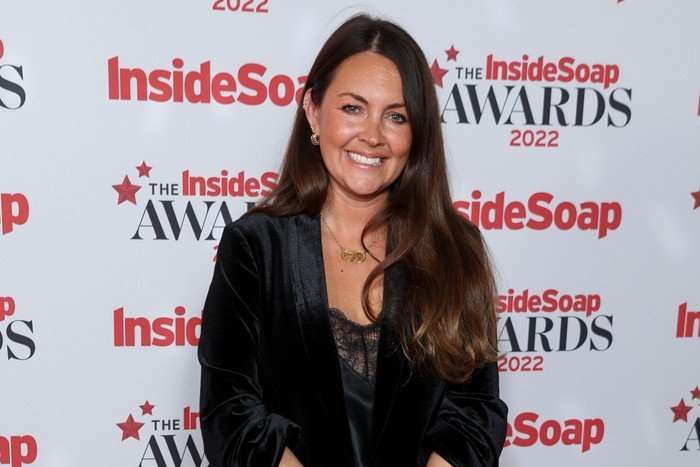 Lacey Turner smiling ahead as she poses for a photo, wearing an all black suit.