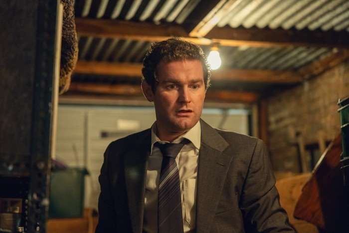Mark Stanley as Ivan Agnew in The Witness standing in a garage, wearing a suit and looking shocked as he looks at something on the ground.