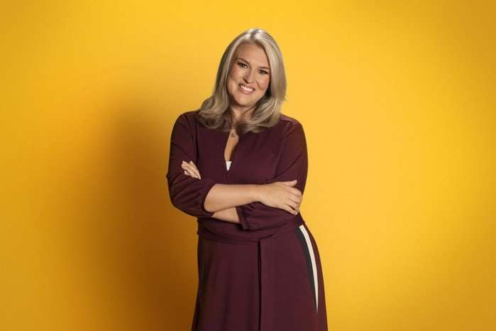 ©ITV Sara Davies stood in front of a mustard yellow background, posing with her arms folded in front of her.