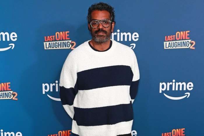 Romesh Ranganathan wearing a white and blue striped jumper, smiling ahead as he poses for a photo.