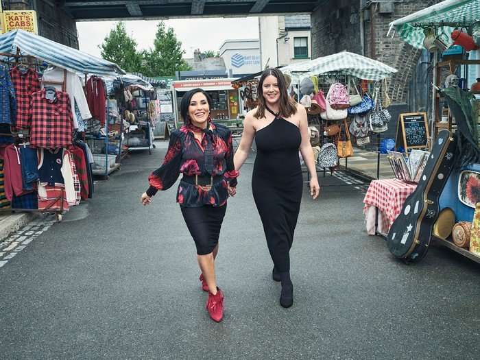 03_20250724_RADIOTIMES_EASTENDERS_194 Jessie Wallace and Michelle Ryan walk hand in hand down the Bridge Street set for EastEnders and both are smiling.