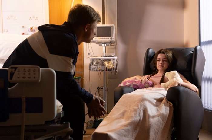 Dylan sits on a bed nearby Betsy in a hospital chair for Coronation Street.