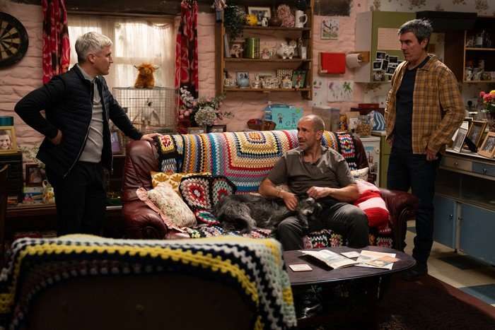 Caleb and Cain stand either side of Sam, who is on the sofa, in a scene from Emmerdale