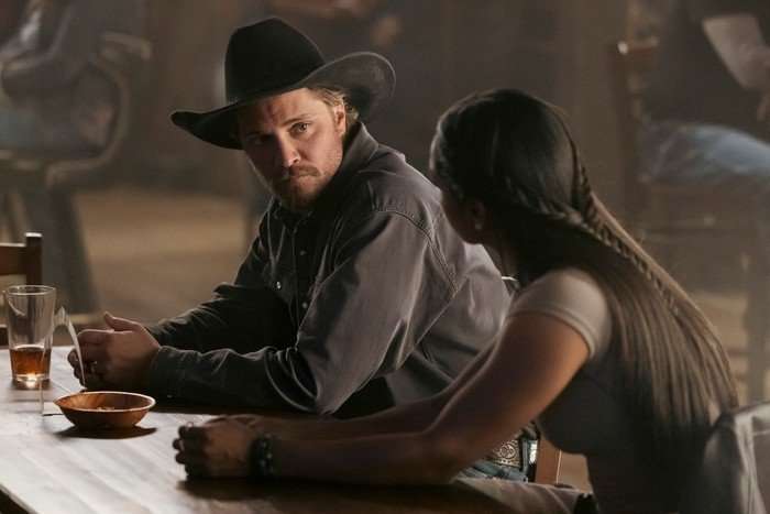 Family Business Luke Grimes as Kayce Dutton in Marshals, sat at a bar, wearing a shirt and a stetson and talking to a woman.