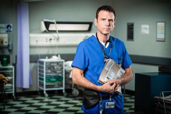 336712,TITLE:Casualty - Portraits 2021 Jason Durr as David Hide in blue scrubs holding a folder in Casualty in 2021.