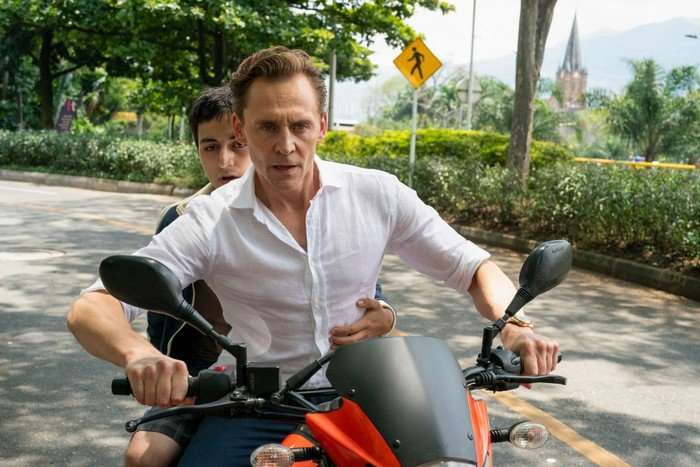 Tom Hiddleston as Jonathan Pine in The Night Manager, riding a motorcycle with a boy on the back of it. Pine is wearing a white, open-necked shirt.