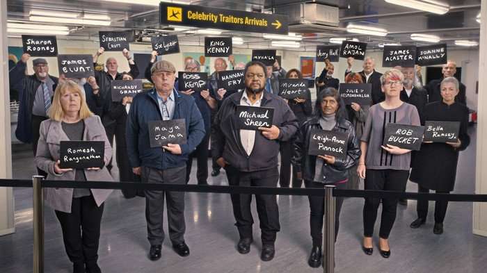 565526,TITLE:The Celebrity Traitors S2 A group of people stood at airport arrivals holding black boards with names on.