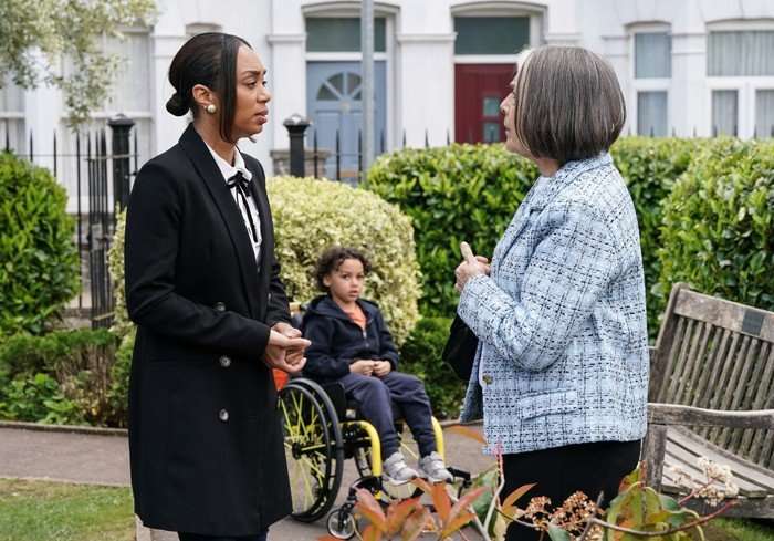 568588,TITLE:Eastenders Sheila and Chelsea chat in the Square, with Jordan in his wheelchair behind in a scene from EastEnders
