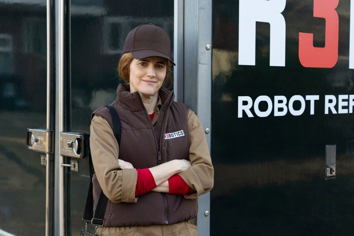 Sarah Kendall as Cass in Ann Droid, wearing a brown top, gilet and cap, and stood by a black van with her arms crossed.