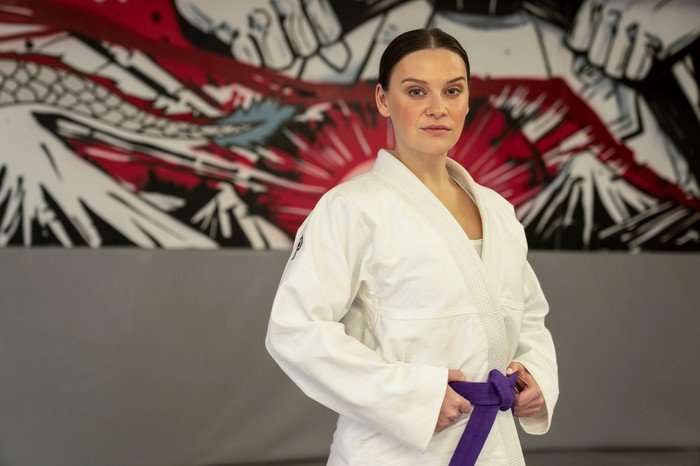 Andrea Valls as Renee in Ann Droid, wearing a white karate outfit, stood in a room and looking at the camera.