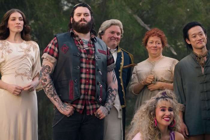 The cast of Ghosts Australia, stood and sat outside together, looking at something off screen.
