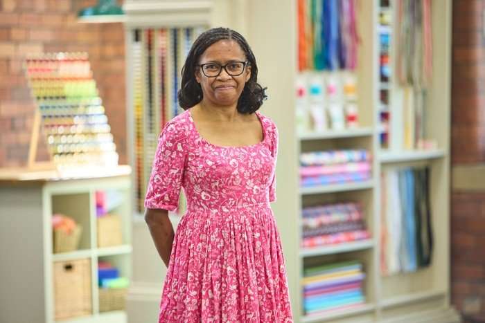 570084,TITLE:The Great British Sewing Bee S12 A woman stood smiling with her arms behind her. She is wearing a pink dress.