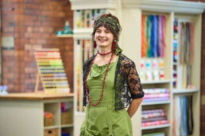 570086,TITLE:The Great British Sewing Bee S12 A woman stood smiling ahead with her arms placed behind her back.