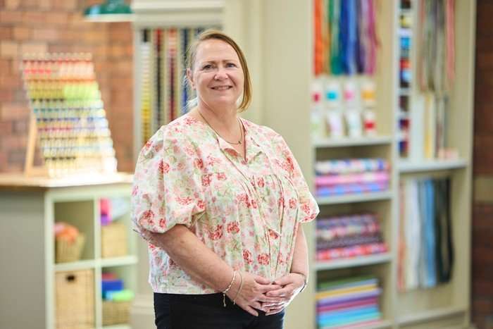 570089,TITLE:The Great British Sewing Bee S12 A woman stood smiling with her arms in front of her, wearing a floral blouse and black trousers.