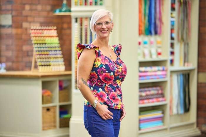 570090,TITLE:The Great British Sewing Bee S12 A woman stood to one side, smiling ahead, wearing a floral blouse and blue trousers.