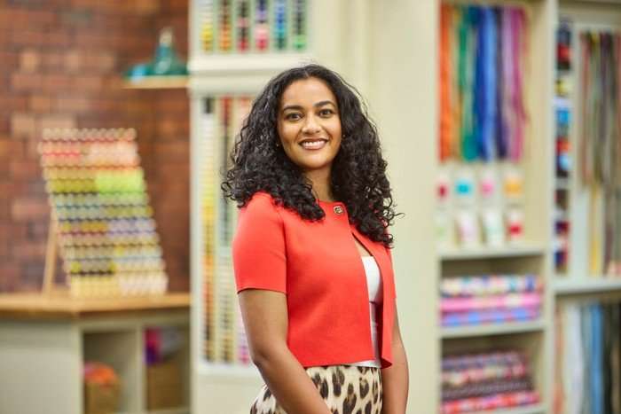 570094,TITLE:The Great British Sewing Bee S12 A woman stood to one side smiling ahead, wearing a white top, leopard print trousers and an orange cardigan.