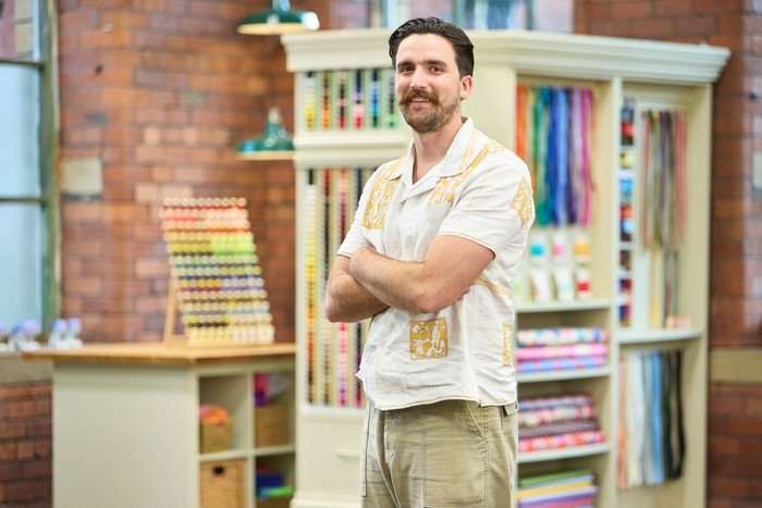 570096,TITLE:The Great British Sewing Bee S12 A man stood to one side smiling ahead with his arms folded in front of his chest.