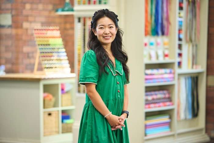 570097,TITLE:The Great British Sewing Bee S12 A woman stood with her hands placed together in front of her, smiling ahead, wearing a green dress.