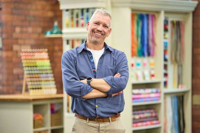570103,TITLE:The Great British Sewing Bee S12 A man stood smiling ahead with his arms folded in front of him. He is wearing a blue shirt and tan trousers.