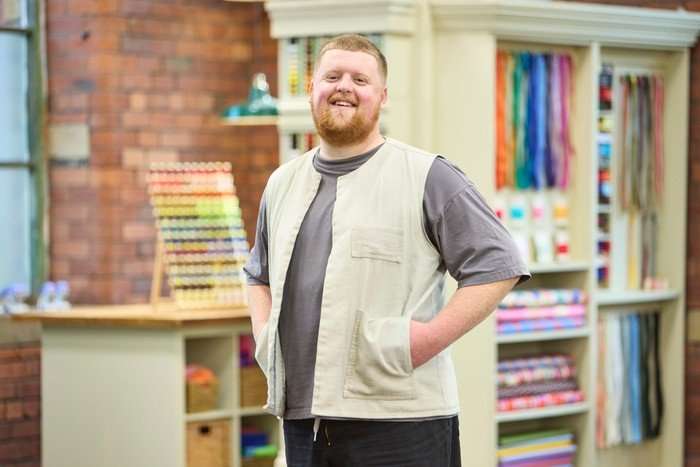 570104,TITLE:The Great British Sewing Bee S12 A man wearing a grey t-shirt, cream gilet and black trousers, smiling ahead with his hands in his pockets.