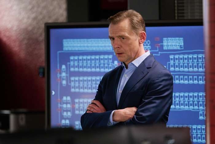 The Undeclared War - Series 2 Ep1 Simon Pegg as Danny Patrick in The Undeclared War, wearing a suit with an open neck shirt and stood in front of a computer screen.