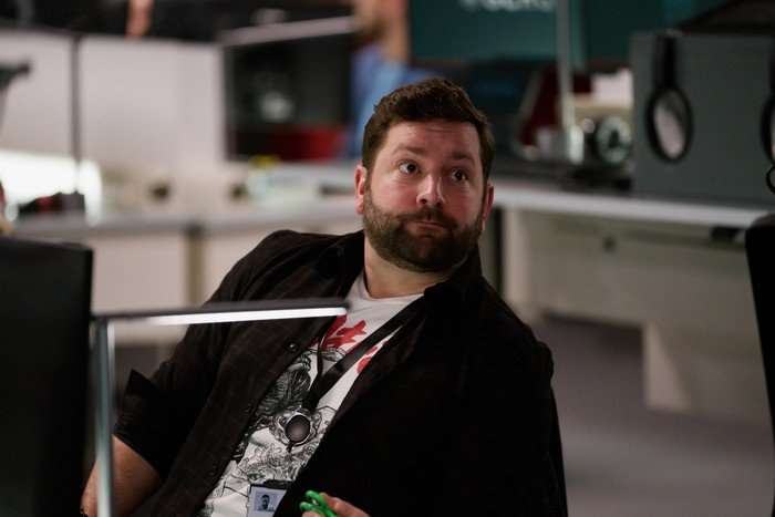 The Undeclared War - Series 2 Ep1 Joss Porter as Phil Draper in The Undeclared War, sat at a desk wearing a white t-shirt and black jacket.