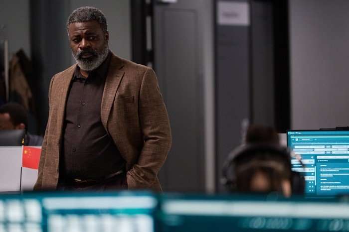 The Undeclared War - Series 2 Ep2 Danny Sapani as Charlie Francis in The Undeclared War, wearing a brown jacket and shirt and stood in an office with his hands in his pockets.