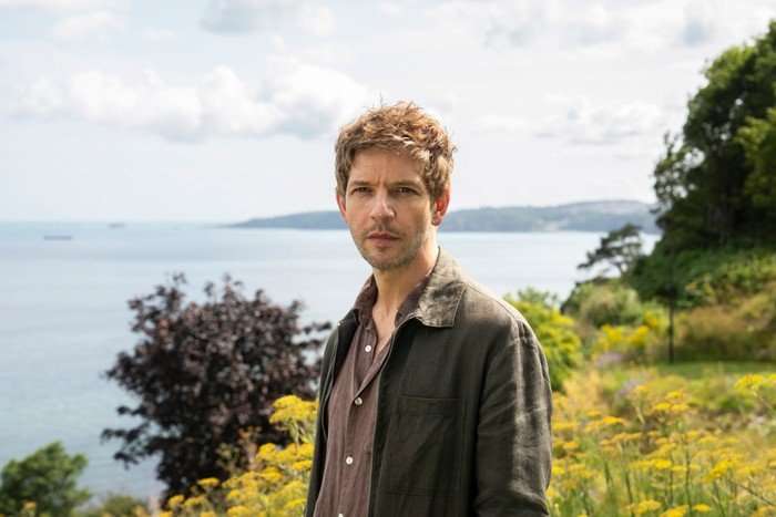 Damien Molony as Jim Bergerac standing in front of a garden and coastal backdrop
