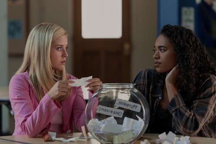 Lexi Minetree as Elle and Chandler Kinney as Kimberly in Elle, sat together, with a fish bowl full of slips of paper in front of them. Elle is holding one of the slips.