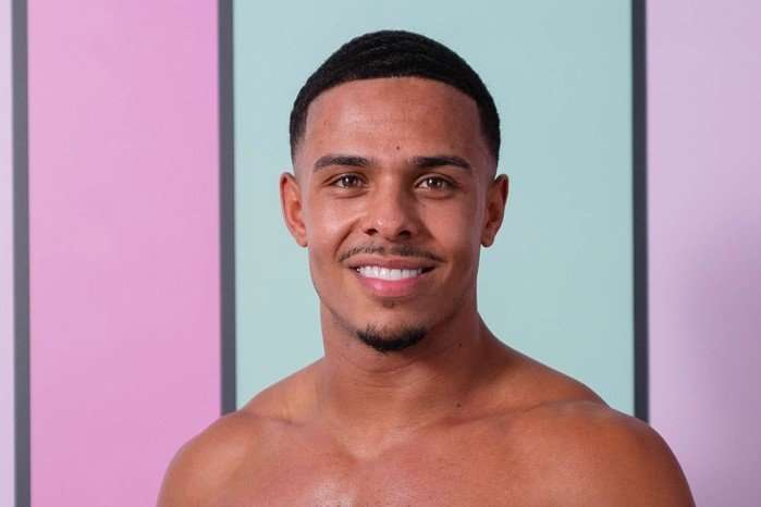 Ethan on Love Island season 13. A close up of a topless man smiling ahead in front of a light blue background.