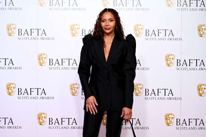 Lois Chimimba, wearing a black outfit and stood in front of a white backdrop with the BAFTA logo on it.
