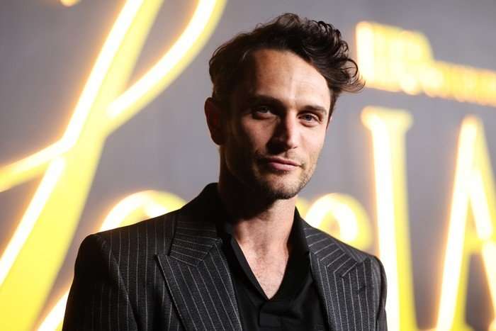 Colin Woodell Colin Woodell poses in a black shirt and grey pinstripe blazer in front of a yellow and grey sign
