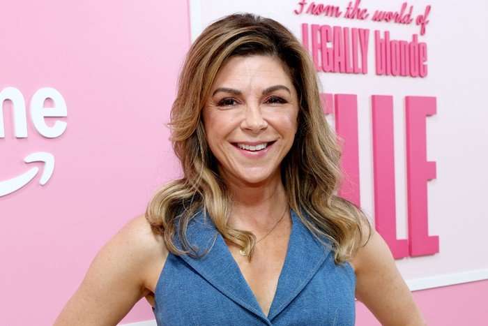 Amy Pietz, wearing a blue dress and stood in front of a pink and white background.