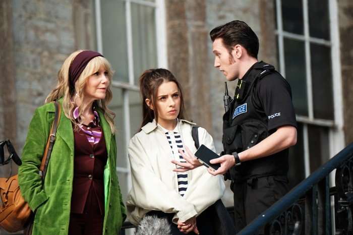 Three people stand outside a stone building in conversation. A police officer in uniform gestures while speaking to two women, one wearing a bright green coat and headband and the other dressed in a light-coloured jacket over a striped top. The younger woman looks concerned as she listens, while the officer appears to be explaining something. The scene takes place on outdoor steps beside large windows, suggesting an ongoing investigation or official inquiry.