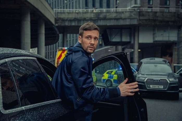 HitPoint-NickBlood Nick Blood stepping out of a car in a police station car park, turning round to look at something.