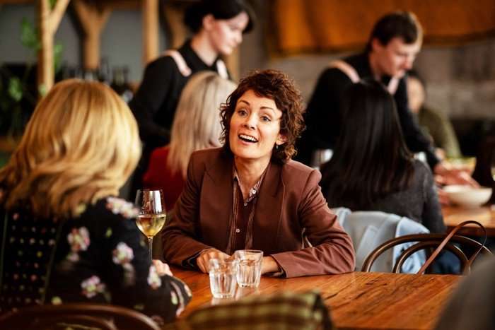 Austin Kate Elliott as Heidi, sat at a restaurant table opposite Ingrid, smiling