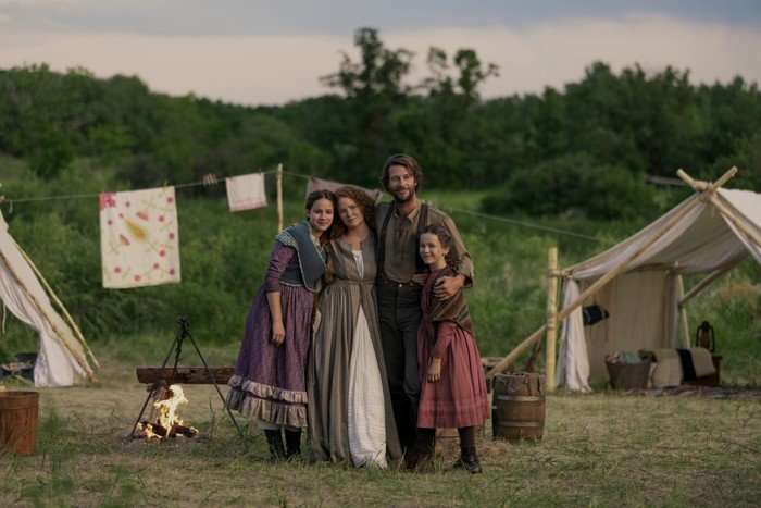 LHOTP_102_Unit_00296RC Skywalker Hughes as Mary Ingalls, Crosby Fitzgerald as Caroline Ingalls, Luke Bracey as Charles Ingalls and Alice Halsey as Laura Ingalls in Little House on the Prairie, stood together in a field, with a clothesline behind them. They are hugging and looking at the camera.