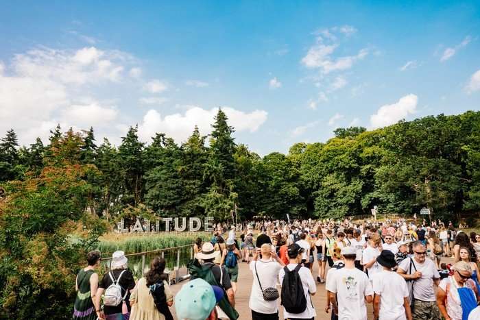Latitude Festival 2025 Crowds walk across a wooden bridge at Latitude Festival, with the large “Latitude” sign, green woodland, and blue summer sky in the background.
