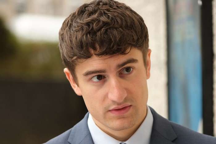 Luke Nunn as Simon Calvert wearing a grey suit