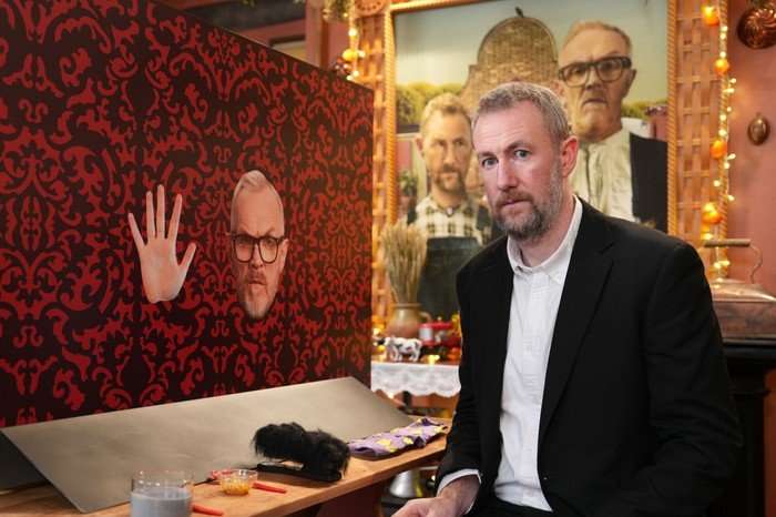 Alex Horne stood next to an image of Greg Davies' face and hand.