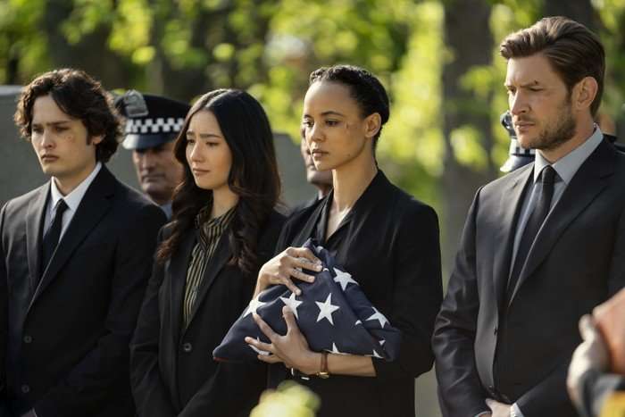 Neagley - First Look Neagley stands at the front of a crowd gathered for a funeral, clutching something wrapped in the United States flag