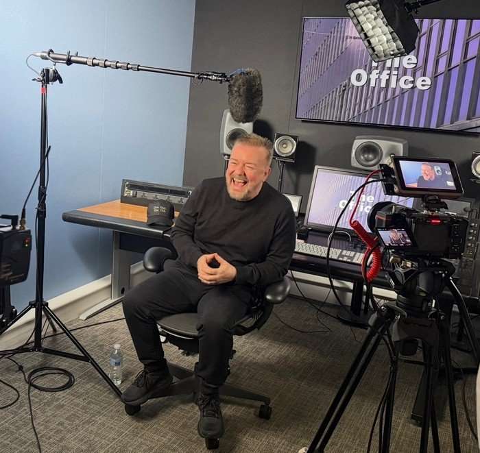 Ricky Gervais sitting in a studio and laughing with a camera filming him and a screen at the back reading 'The Office'.